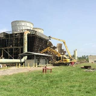 A construction site with a large yellow excavator, operated by a reputable demolition company, working near a partially demolished industrial structure amidst green grassy surroundings.