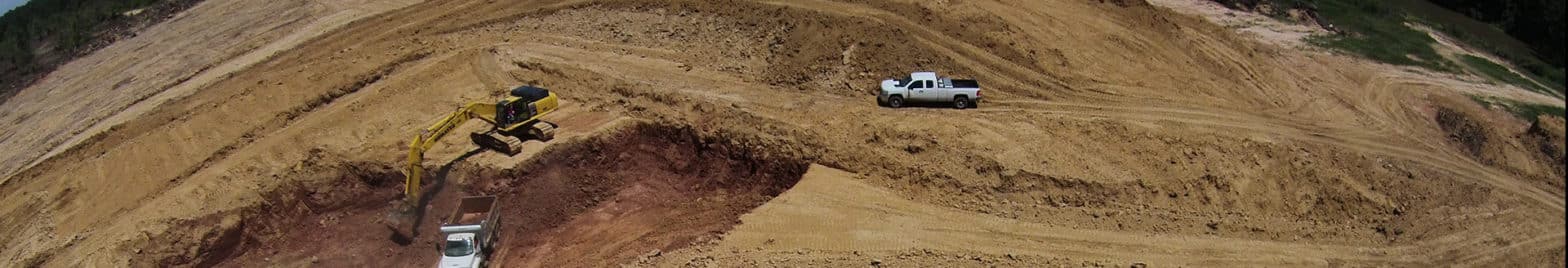 Aerial view of a construction site with an excavator loading a truck, while a white vehicle from the environmental consulting team is parked on a dirt road.
