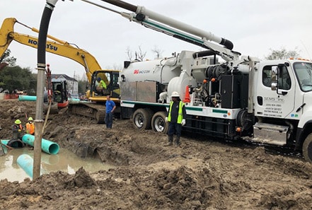 Construction workers from a remediation company are operating heavy machinery to install large pipes in a muddy construction site. An excavator and a vacuum truck are actively being used while ensuring industrial hygiene services are maintained throughout the process.