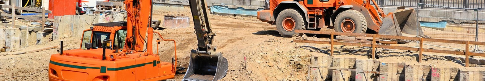 Two orange excavators are operating on a construction site, managed by an industrial services company. One excavator is digging with its arm, while the other is driving near a wooden barrier. The ground appears to be mostly sand and dirt.