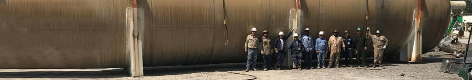 A group of 11 people wearing hard hats and protective clothing stands in front of a large industrial cylindrical structure, representing an industrial services company at the worksite.
