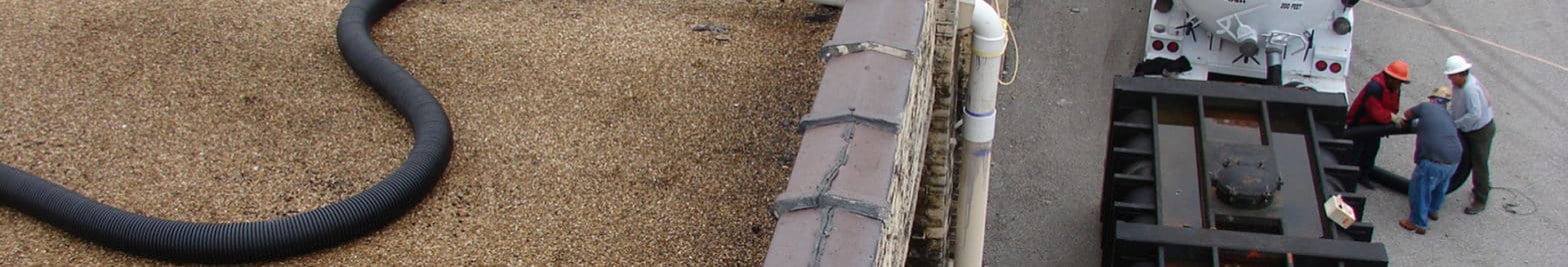A rooftop gravel vacuuming system is in use, with a large black hose and vacuum truck visible. Three workers in hard hats from a remediation company oversee the operation from the ground below.