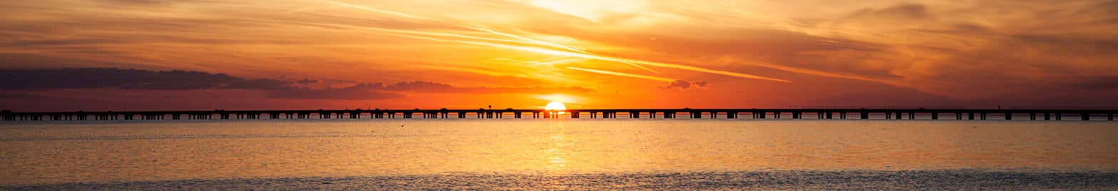 A long pier stretches out over calm water at sunset, with an orange and yellow sky reflecting on the surface, embodying the same serene focus and precision an industrial services company brings to its work.
