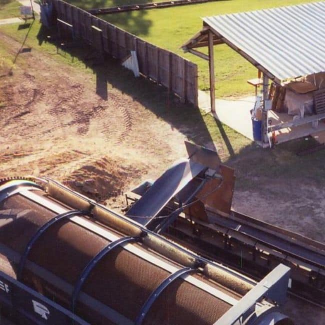 An outdoor piece of industrial machinery, likely used for sorting or processing materials, is positioned near a small shed with a metal roof and a fenced area, illustrating the expertise of an industrial services company.