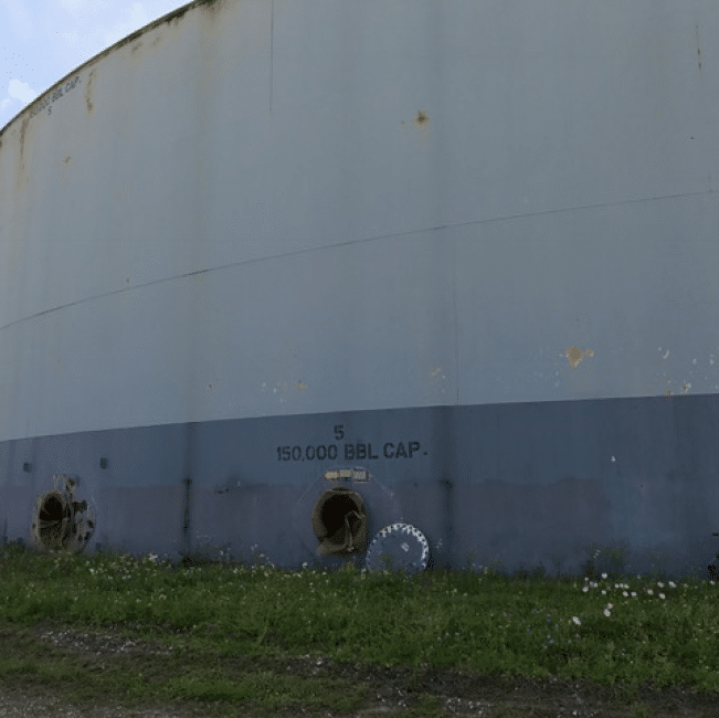 A large cylindrical industrial storage tank labeled "5 150,000 BBL CAP" with rusting areas near the base stands on grass under an overcast sky, highlighting the urgent need for remediation services from an industrial services company.