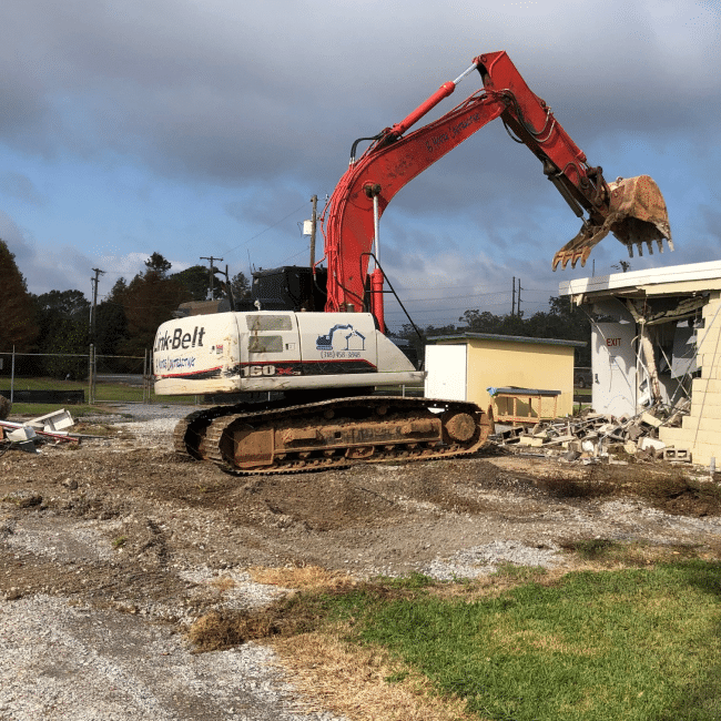 A large excavator from a top-tier demolition company demolishes a small building. The scene includes debris around the site and power lines in the background, showcasing their comprehensive industrial services.