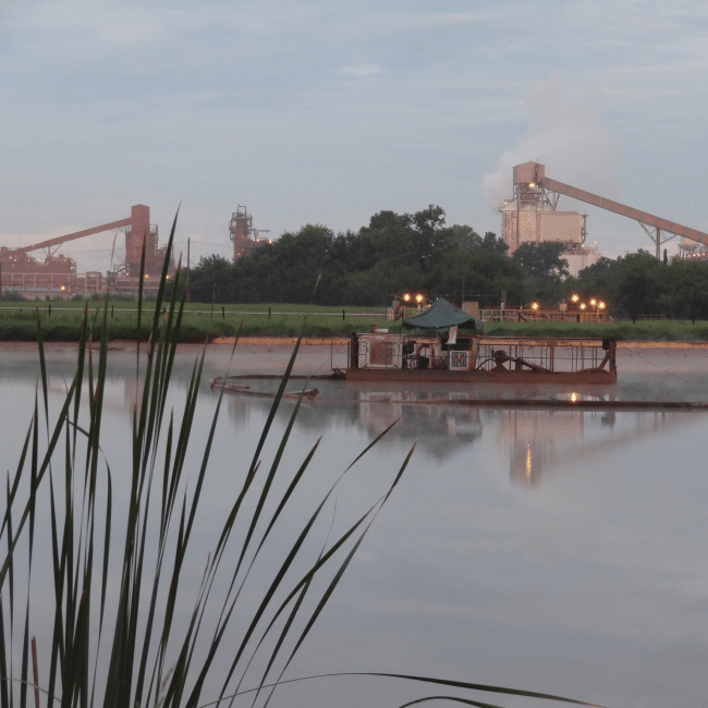 A barge with lights is floating on a calm river, with an industrial services company’s factory and smokestacks emitting smoke in the background under a cloudy sky. Tall grass is visible in the foreground.