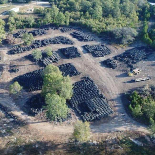 An aerial view of a large open area with numerous stacks of black tires arranged in rows, surrounded by trees and vegetation, possibly overseen by an industrial services company ensuring proper organization and safety.