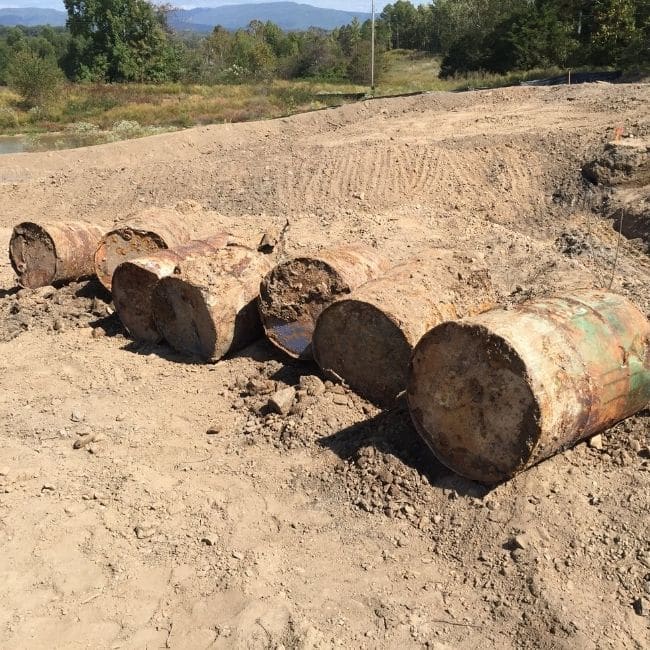 Six rusted metal barrels partially buried in dirt at a construction site with trees and hills in the background, ideal for an environmental consulting company to assess.