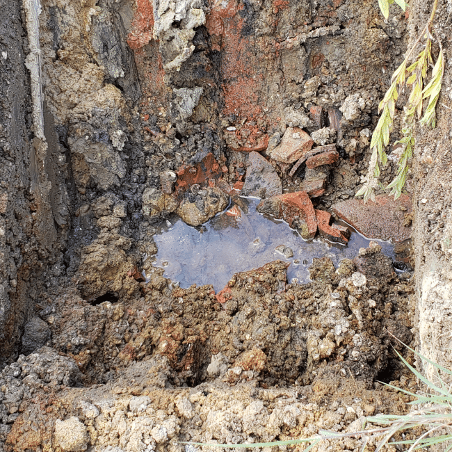 A hole in the ground filled with water and broken pieces of reddish material, surrounded by soil and rocks from a recent demolition company project. A plant with green leaves is partially visible on the right side.