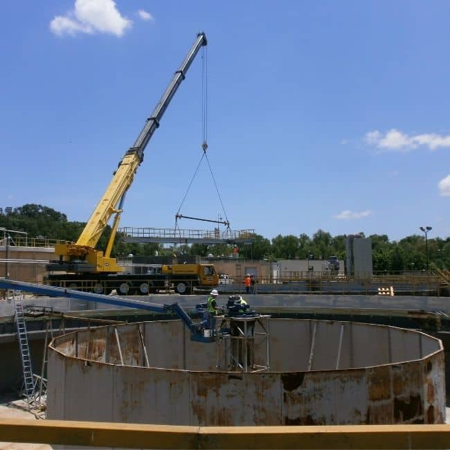 A yellow crane from an industrial services company lifts a large metal structure at a construction site with circular concrete forms. Workers in safety gear are on-site performing tasks under the clear blue sky.