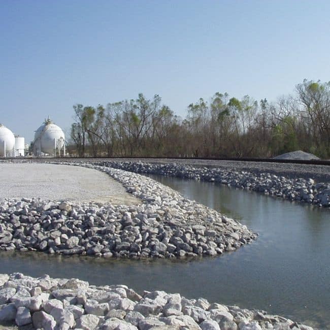 A landscape view of an industrial site with two large spherical storage tanks, a channel of water lined with rocks, and trees in the background under a clear blue sky, highlighting the work of an environmental consulting company ensuring sustainability.