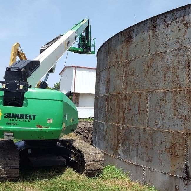 A green Sunbelt Rentals lift is positioned beside a rusted metal silo, with a worker in a green vest standing on the lift's platform near the top of the silo, marking the precision of an industrial services company at work.
