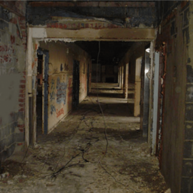 A dark, abandoned hallway with peeling paint, exposed wires, and graffiti on the walls. The floor is strewn with debris and the space appears neglected and dilapidated, prime for an industrial services company’s expertise to transform it.