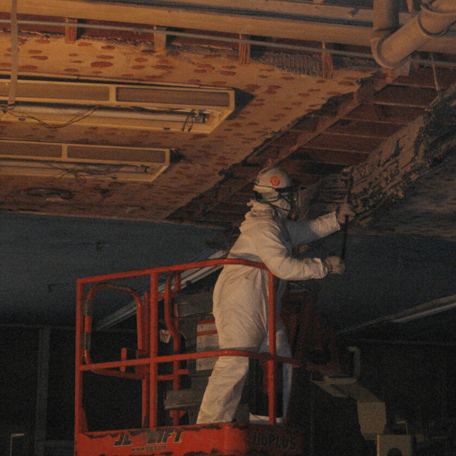 A worker in protective gear stands on an orange lift platform, using a tool to remove damaged materials from a ceiling, showcasing the expertise of a demolition company.