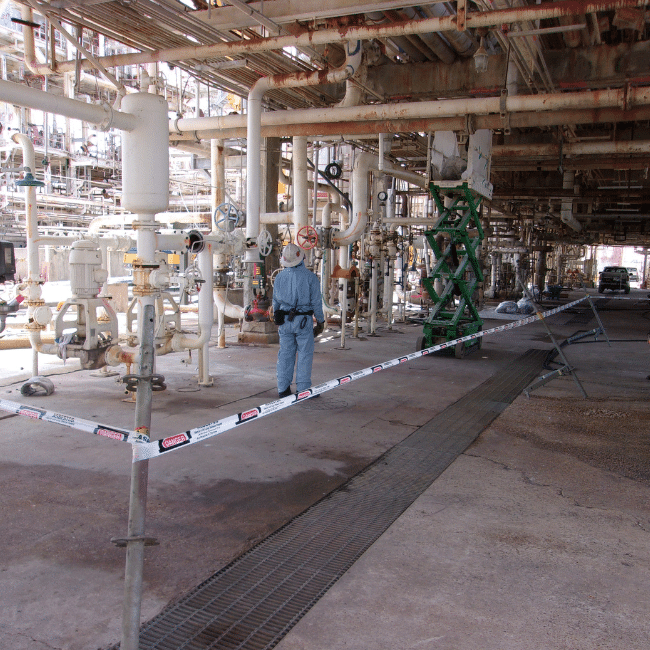A worker in blue protective clothing examines pipes and valves in an industrial facility managed by an industrial services company. The area is cordoned off with caution tape, and a green scaffolding is visible.