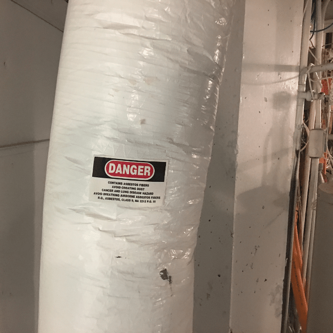 A large pipe wrapped in white material with a red and black "Danger" sign warns of asbestos fibers, instructing to avoid breathing dust to prevent disease hazards. This is where industrial hygiene services play a crucial role, ensuring safety and health standards are met.