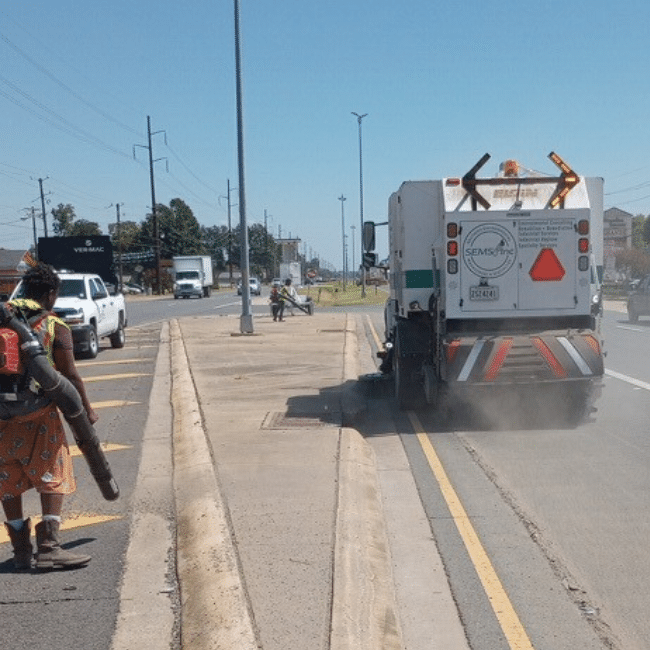 A person operates a leaf blower on a median strip while an industrial services company's street sweeper vehicle cleans the adjacent road.