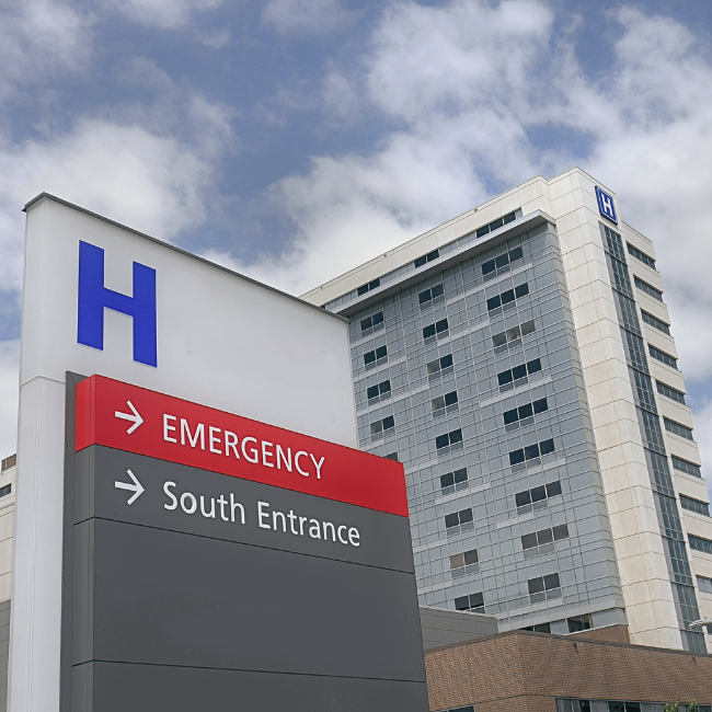 Hospital building with a large "H" sign. Directional sign indicates Emergency to the left and South Entrance straight ahead. Nearby, an industrial services company ensures safety through meticulous industrial hygiene services for all visitors and staff.