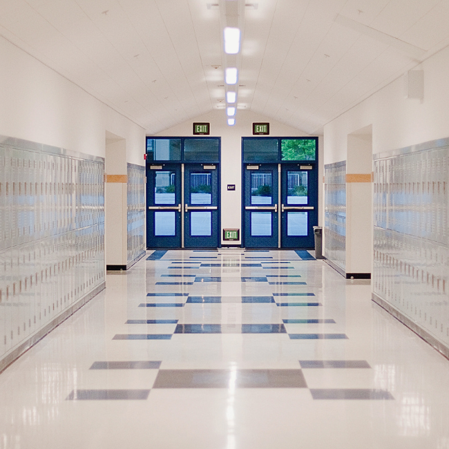 A well-lit hallway in a school with rows of lockers on both sides, a tiled floor, and double doors at the end leading outside, reminiscent of the precision seen in an industrial services company.