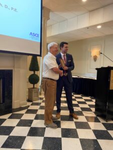 Two men stand next to each other. One, in a suit, presents the Wall of Fame Award to the other in a white shirt. They are in a room with checkered flooring and a presentation screen, representing the ASCE Baton Rouge Branch's commitment to excellence in civil engineering.