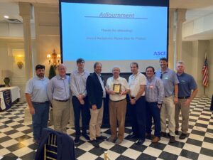 A group of men stands in front of a presentation screen displaying "Adjournment." One man, honored with the Wall of Fame Award from the ASCE Baton Rouge Branch, proudly holds a plaque as eight others stand next to him.