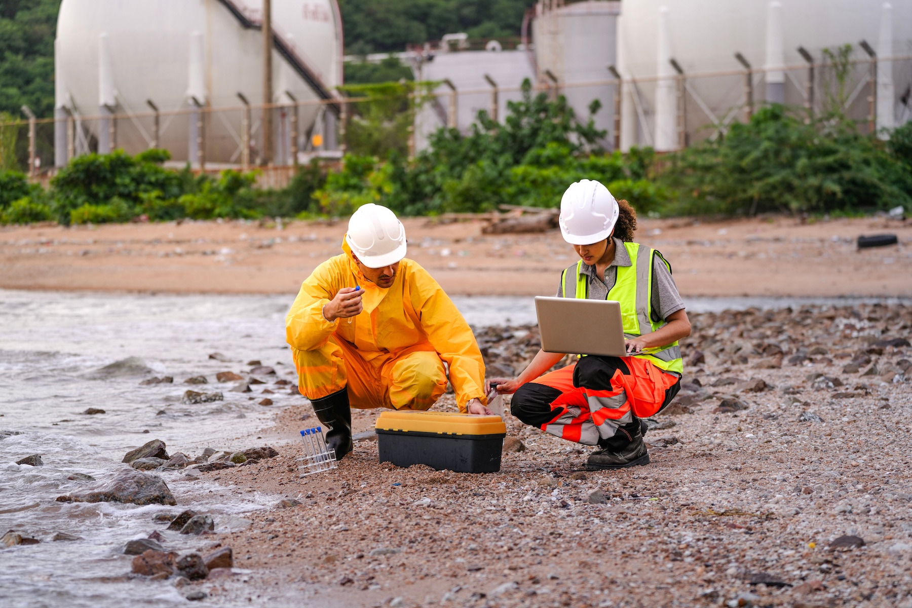 Two environmental engineers in safety gear collecting soil samples near gas storage tanks.